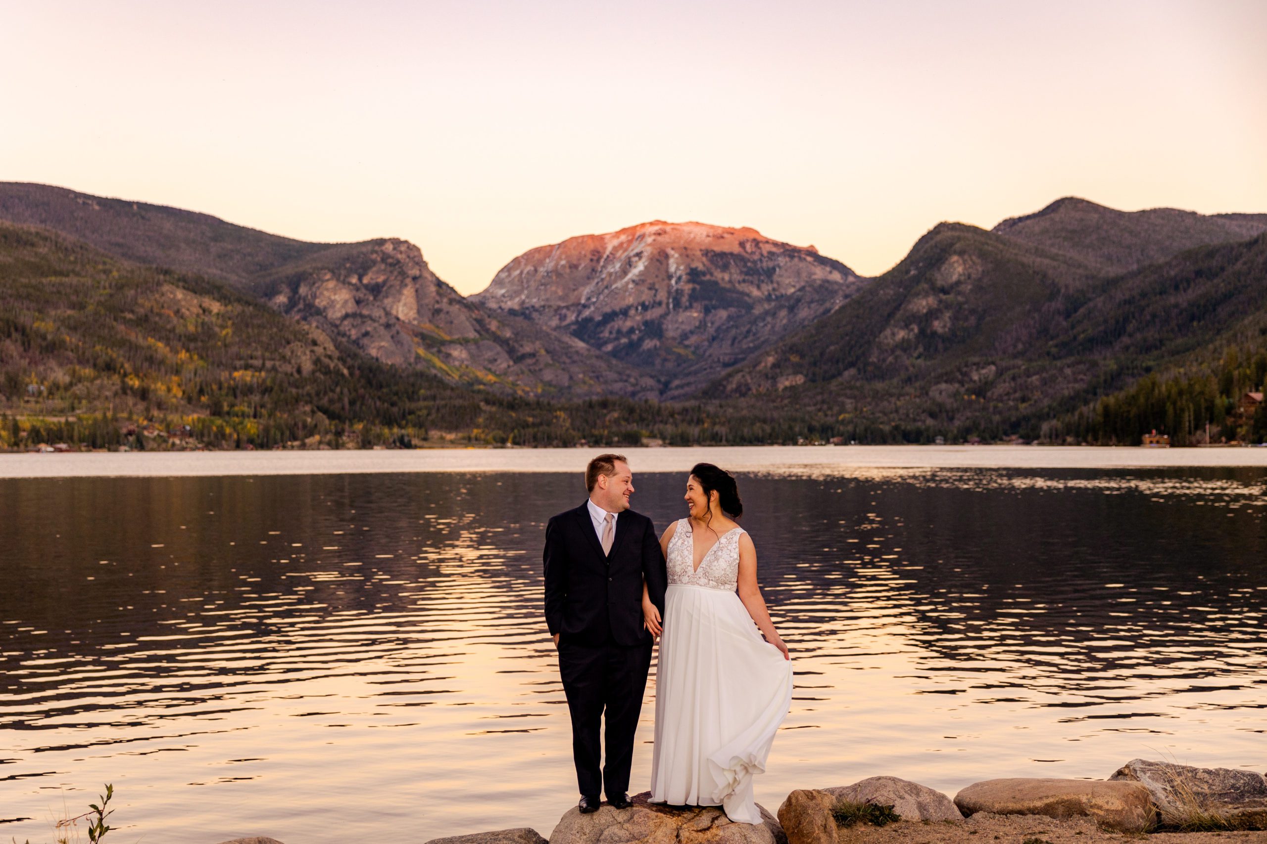 Bride and groom standing on the shore of Grand Lake with Shadow Mountain as a backdrop smiling at each other