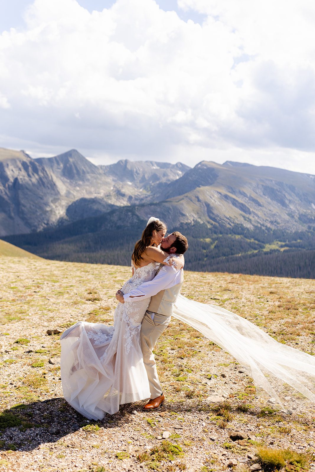 The groom holds his bride as they kiss on the mountain top. 