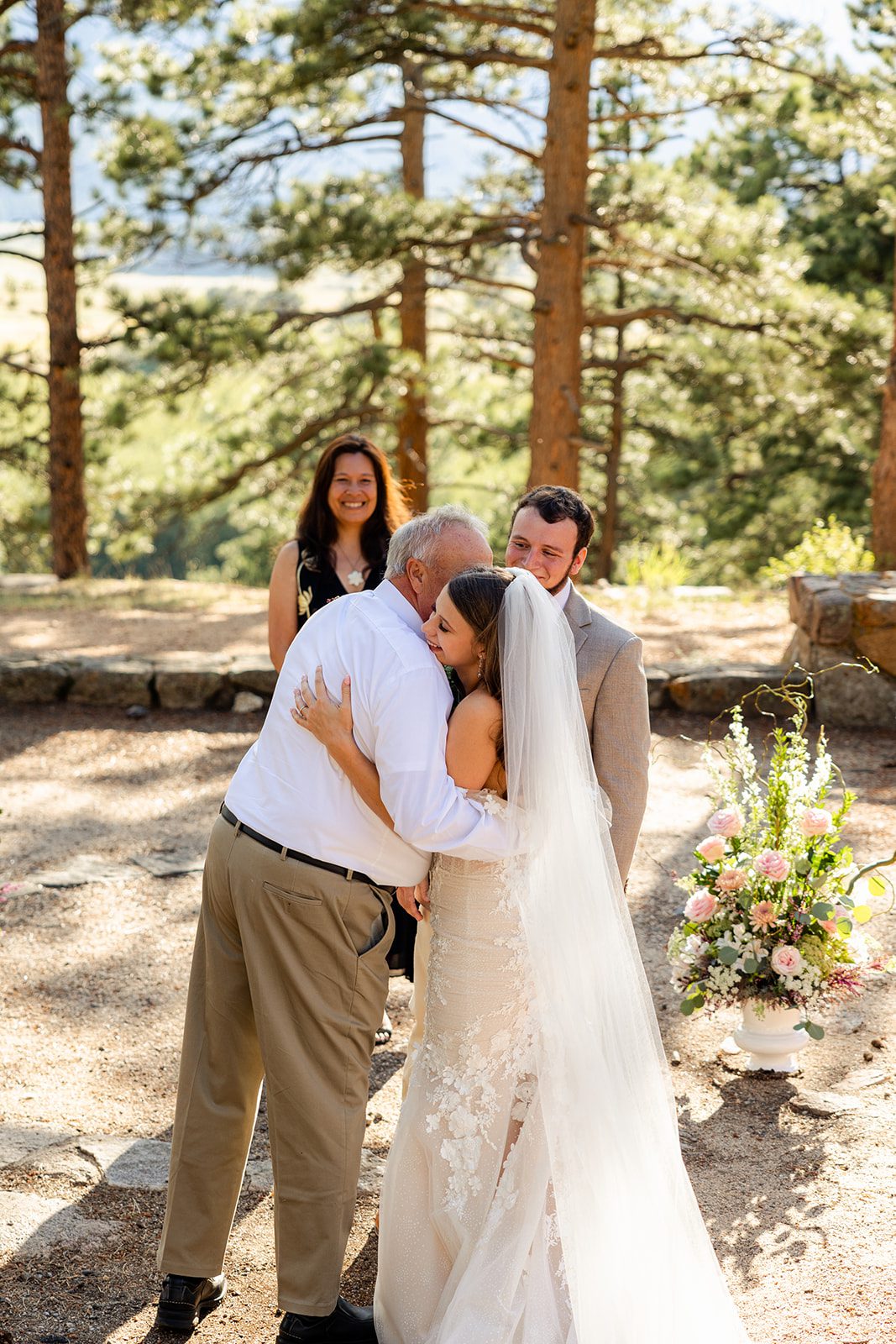 The father of the bride hugs his daughter, the bride. The groom looks at them with love in his eyes. 