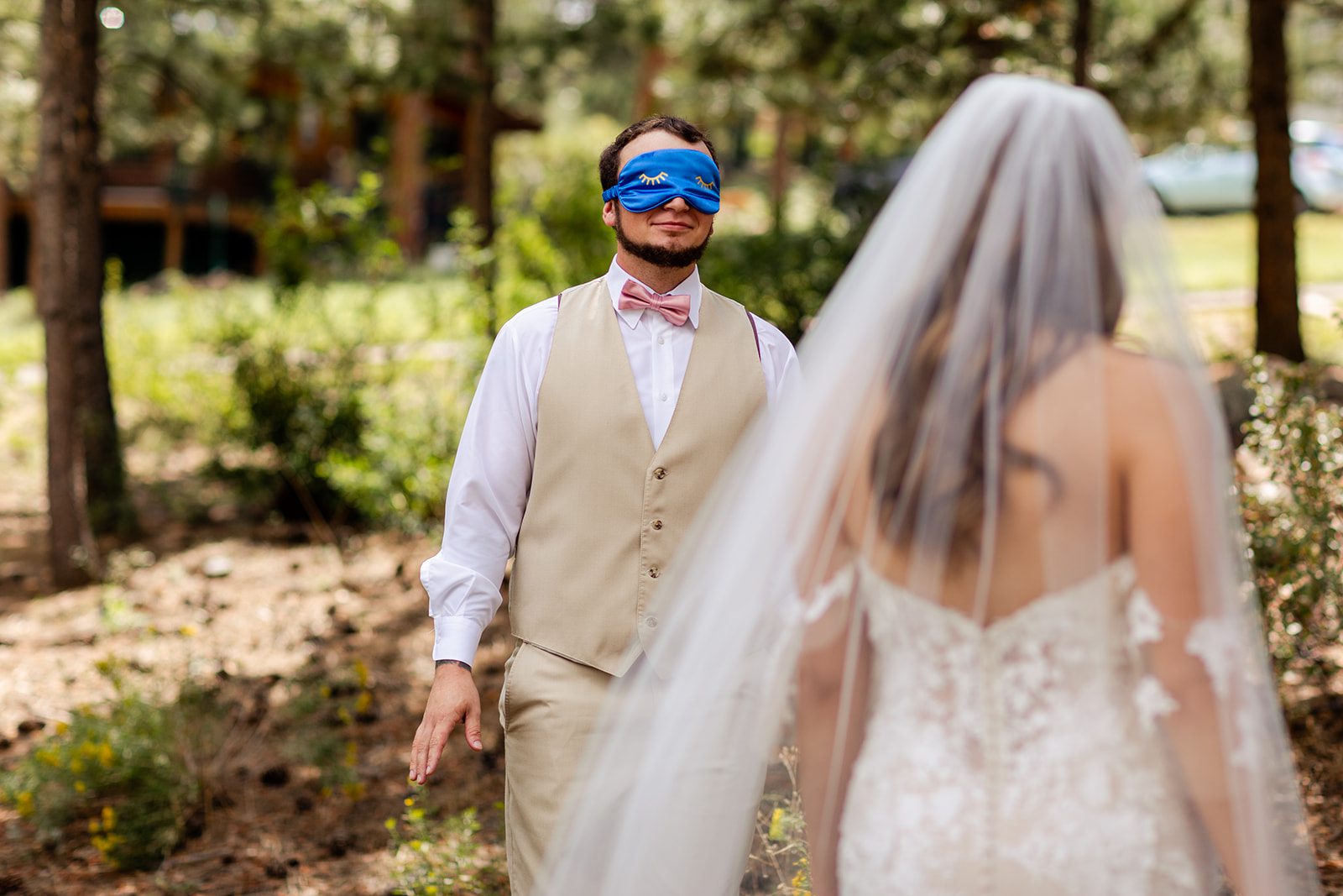 The groom turns to see his bride for the first time on their wedding day. His eyes are still covered with a a blue eye mask with gold eye lashes. 