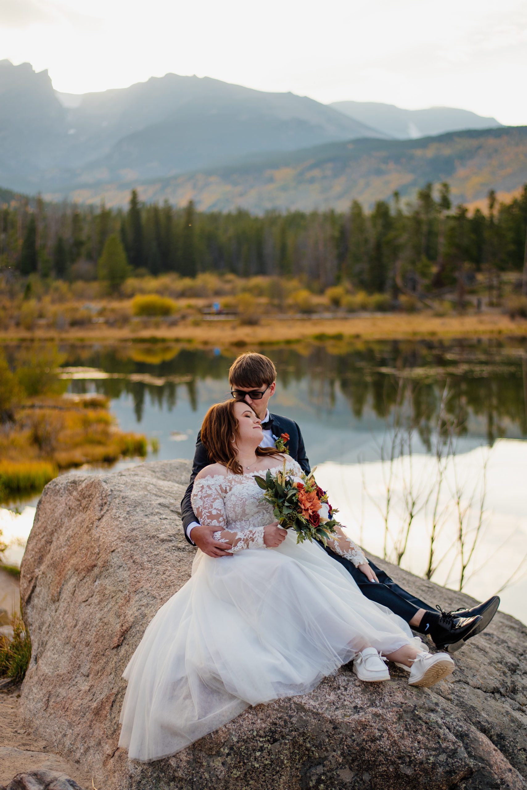 Groom kisses bride's forhead as they are sitting on the rock at Sprague Lake - RMNP