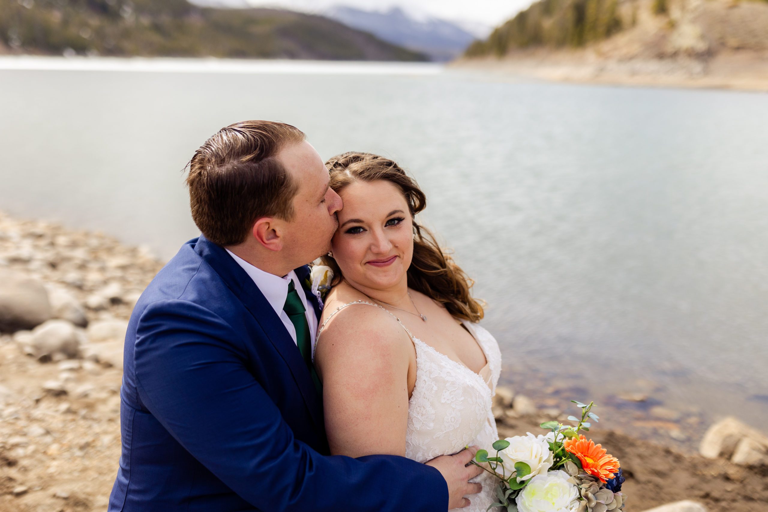 the beautiful bride smiles at the camera while her husband kisses her on the forehead, before their Sapphire Point Elopement ceremony. 