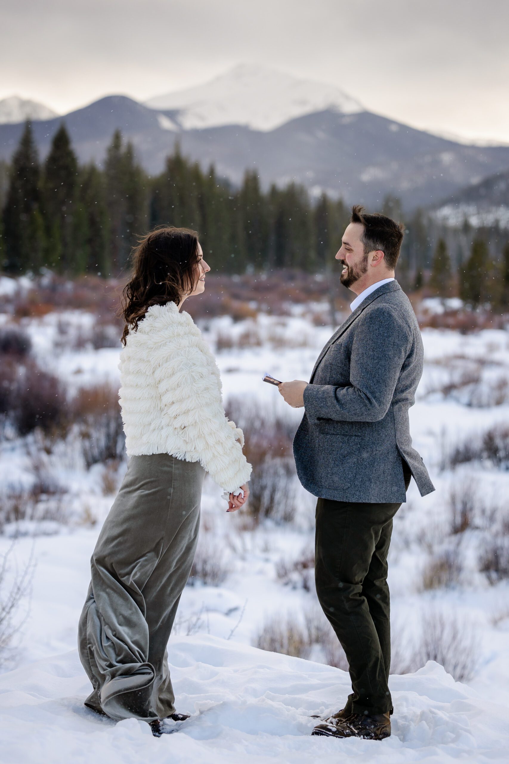 the bride and groom smiling at each other, the snow is falling all around them, the gorgeous white-capped mountain in the distance at their Winter Park elopement ceremony. 
