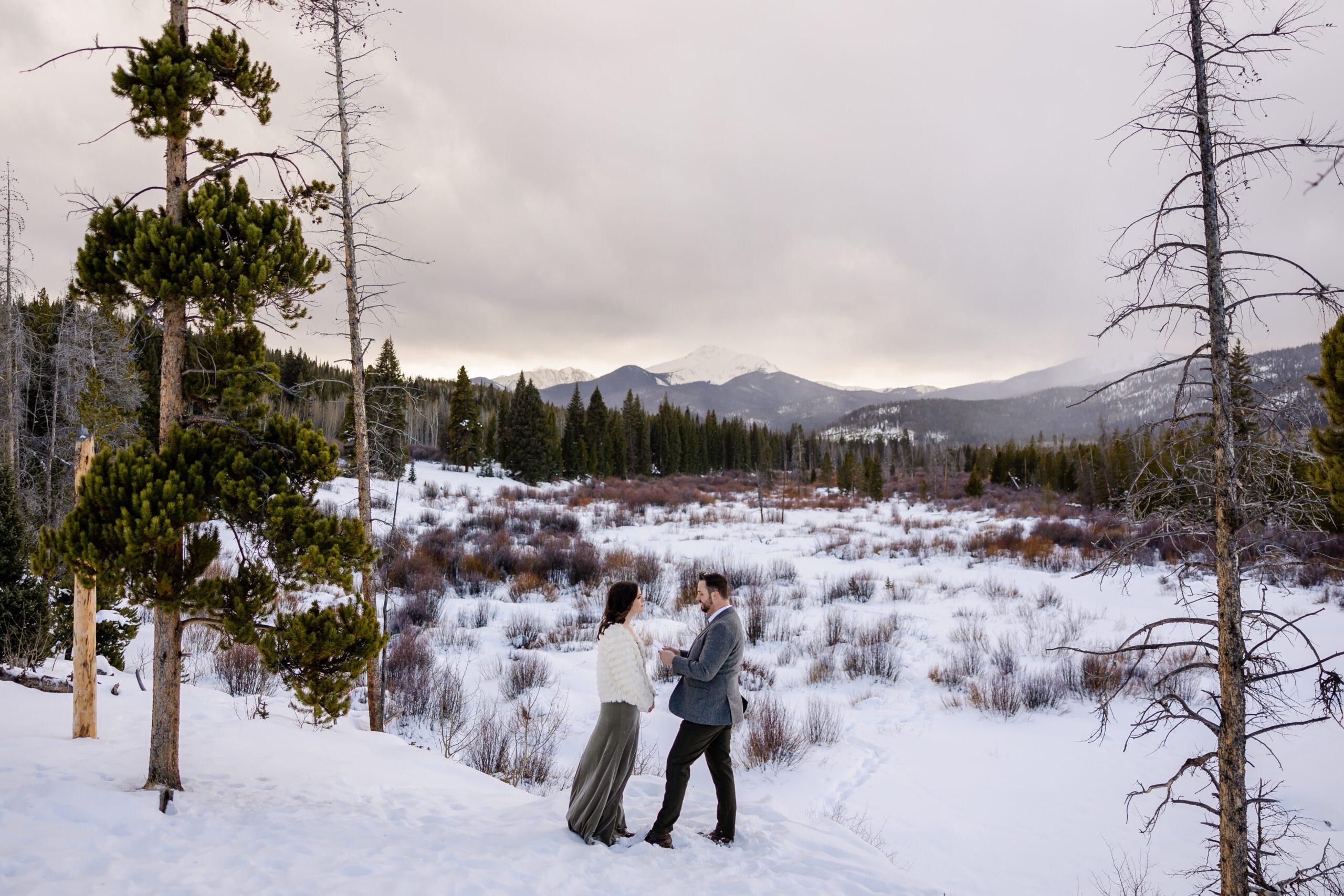 the bride and groom stand in the snow, snow-capped mountains in the distance at their Winter Park elopement ceremony. 