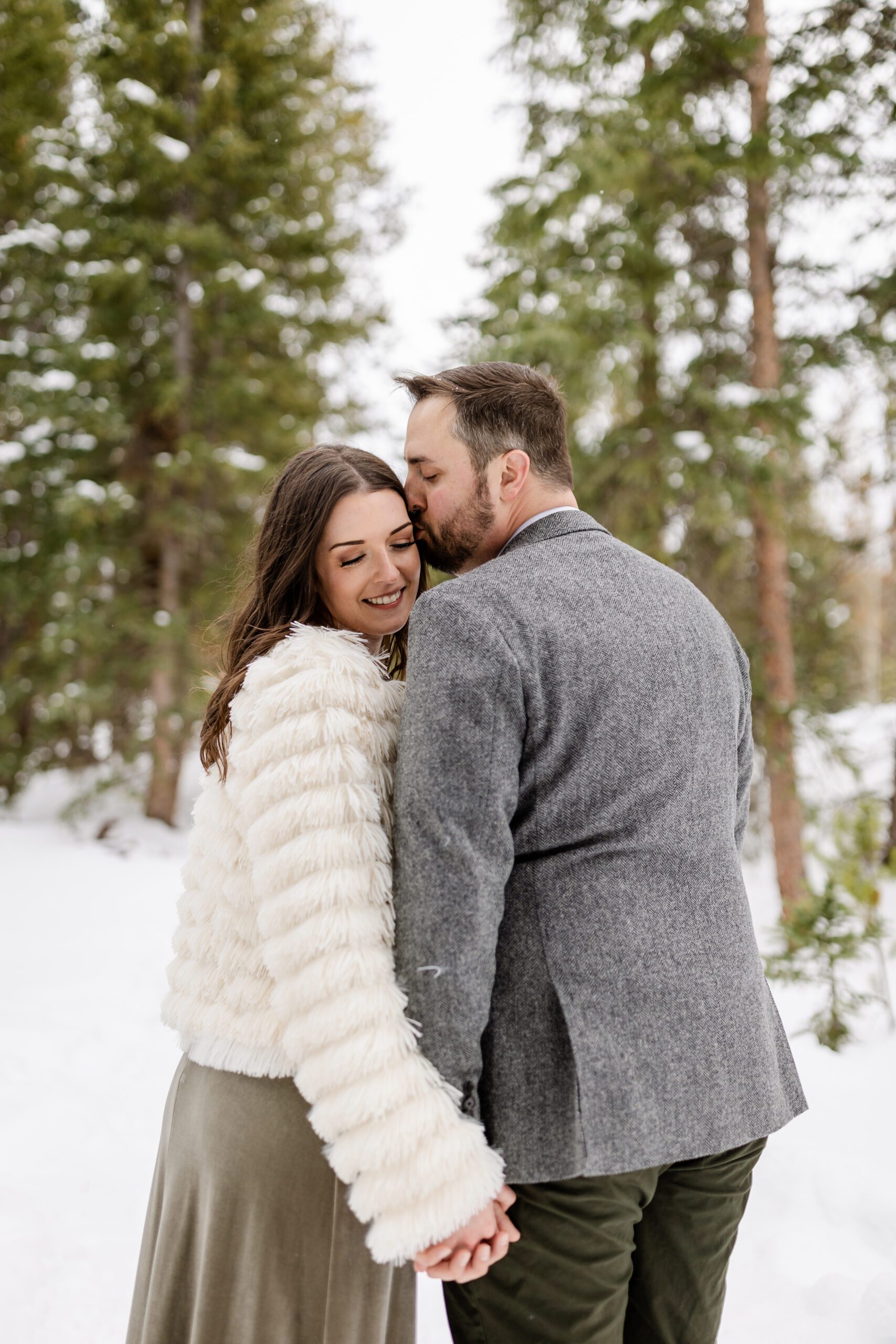 bride smiles sweetly, wearing a white fur coat, while her groom kisses her cheek at their Winter Park elopement.