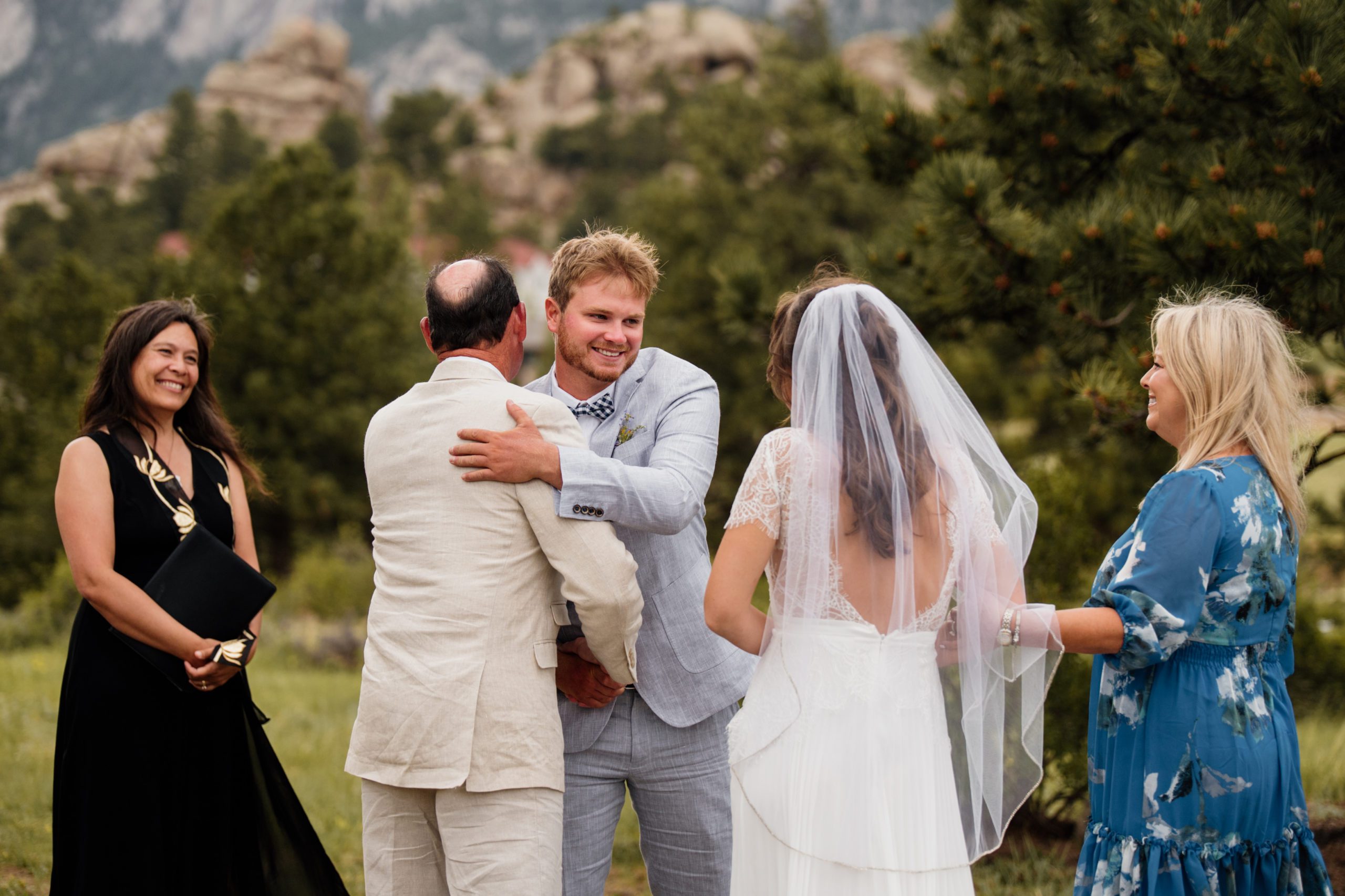 The groom hugs his bride's dad at the end of the aisle before their ceremony at The bride walks down the path towards her groom at their Knoll Willows elopement in Estes Park.