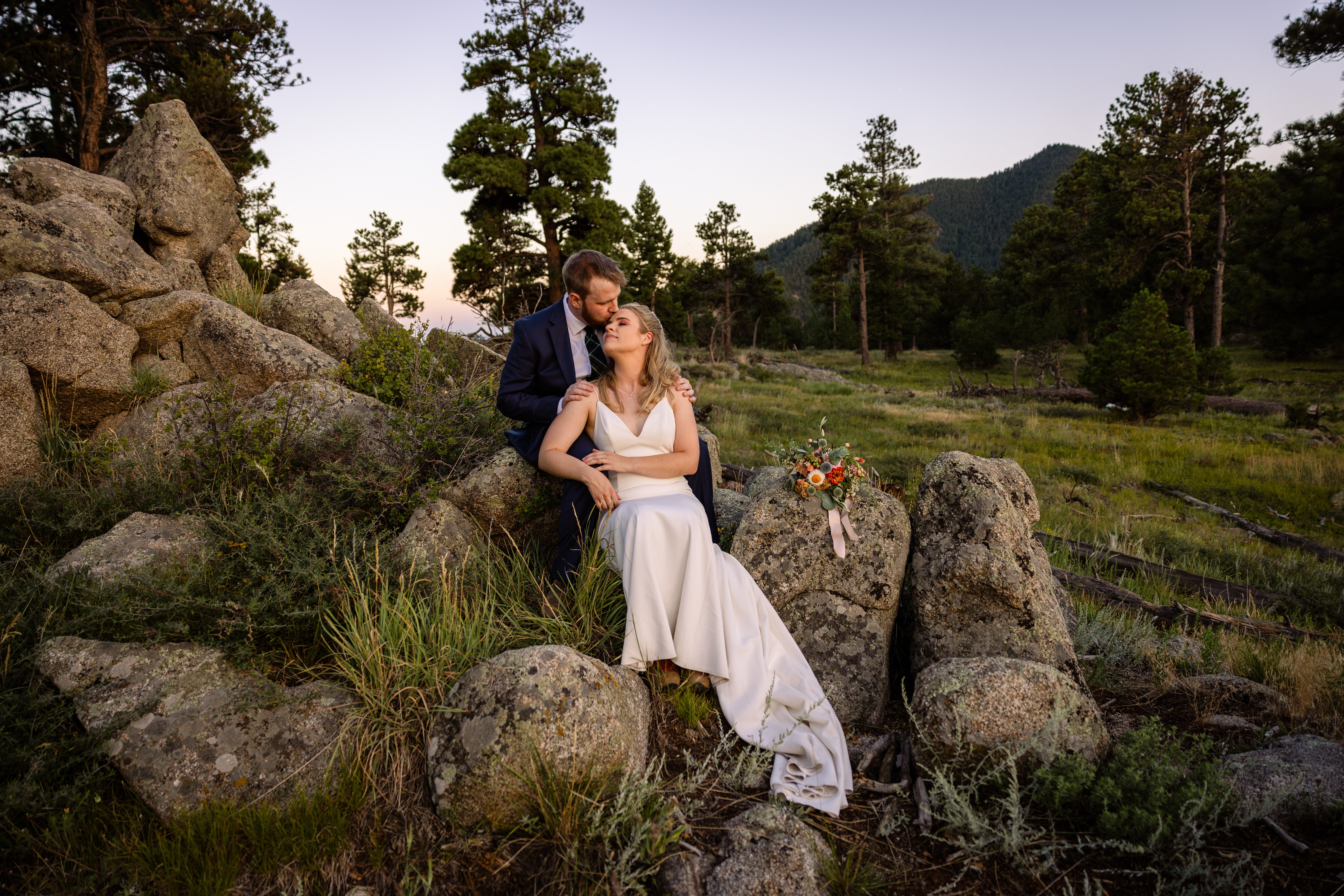 The groom kissing his bride on the forehead while they sit on a rock at sunset after their Sunrise Amphitheater wedding.