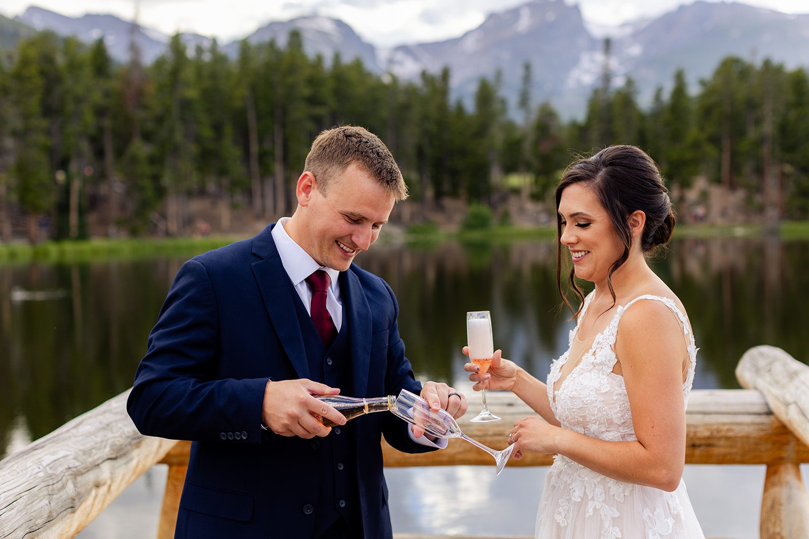 Groom pours champagne for his bride in front of Sprague Lake after their summer elopement