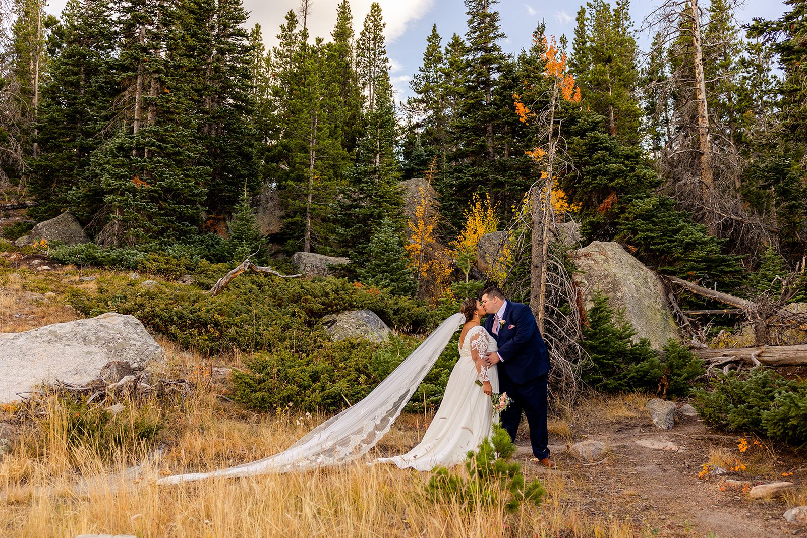 bride in a gorgeous white wedding dress with long veil, kissing groom in Rocky Mountain National Park for their fall elopement at Sprague Lake. 